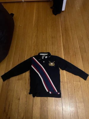 Mörkblå långärmad pikétröja Northwoods - Mörkblå långärmad pikétröja från Northwoods med broderad guldfärgad logga och texten 'Protect & Defend 1986' på bröstet. Snygga diagonala ränder i rött, vitt och blått framtill. Klassisk krage med knappar och sportig stil.