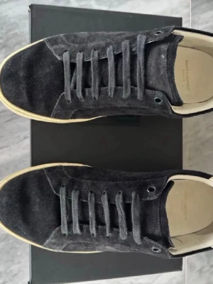 Svarta Saint Laurent sneakers i mocka - Snygga svarta sneakers från Saint Laurent i mjuk mocka med klassisk låg profil och beige sula. Skorna har svarta skosnören och diskret branding på innersulan och yttersulan. Perfekta för en stilren och trendig look. Priset kan diskuteras vid snabb affär.