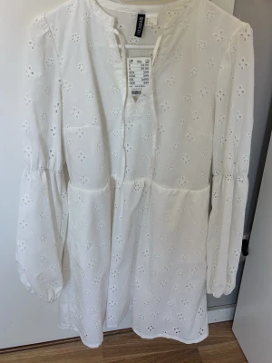 Vit broderad klänning från Hm - Söt vit klänning från Hm med broderade blommor och långärmade puffärmar. Klänningen har en avslappnad passform, rund halsringning och är tillverkad i bomull. Perfekt för vår och sommar!