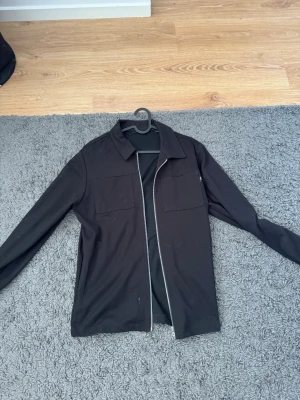 Svart overshirt med dragkedja - Stilren svart overshirt med dragkedja framtill och två bröstfickor. Overshirten har klassisk krage och raka ärmar. Perfekt att bära öppen eller stängd för en avslappnad look. Materialet ser ut att vara mjukt och följsamt.