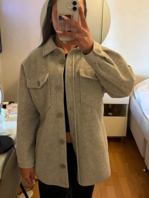 Ljusgrå kappa med fickor - Stilren ljusgrå kappa med stora bröstfickor och knappar framtill. Klassisk krage och är tillverkad i ett mjukt, tjockare material som ger en avslappnad look. Perfekt att bära öppen eller stängd över en t-shirt.