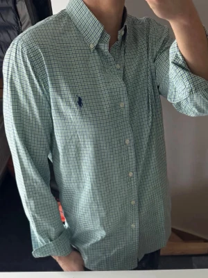 Grön rutig skjorta från Ralph Lauren - Klassisk grön och vit rutig skjorta från Ralph Lauren med button-down krage och blå logga broderad på bröstet. Skjortan har långa ärmar, vita knappar och är tillverkad i mjuk bomull. Perfekt för en clean och stilren look.
