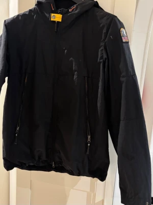 Svart vindjacka Parajumpers Smash - Svart vindjacka från Parajumpers, modell Smash. Jackan har huva. Parajumpers UTA Jacket – L – Ny med lappar  Helt ny svart vind jacka med hoodie Parajumpers jacka (modell UTA) i storlek L.   Aldrig använd – endast provad. Alla lappar kvar. Felköp av mig. Inköpt på Thernlunds.   Klassisk svart modell med PJS-patch på armen och orange meshfoder. Certilogo finns för äkthetskontroll.  Nypris ca 4999 kr Säljes för 3000 kr 