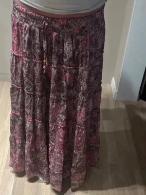 Långkjol designbysi - Långkjol med blommigt mönster i rosa och lila toner. Kjolen har resår i midjan och snörning med träpärlor som detalj. Tillverkad i ett lätt och luftigt material, perfekt för varmare dagar. Jag är 168 och på bilderna ser man hur den sitter på mig, kommer inte till användning trots att den bara används en gång och säljs därför. Storlek Xs-S