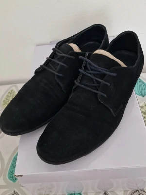 Svarta mockaloafers med snörning - Säljer ett par eleganta derby-skor från HUGO BOSS i svart kalvmocka. En klassisk sko som passar perfekt till både kostym och jeans. ​Storlek: 42 (UK 8 / US 9). ​Material: 100% kalvläder (mocka) med gummisula. ​Skick: Fint använt skick. Se bilder för detaljer på sula och material. ​Tillbehör: Originalkartong medföljer. ​