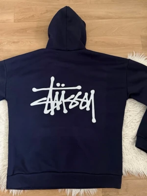 Mörkblå Stüssy hoodie med logga - Snygg mörkblå hoodie från Stüssy med stor vit logga i graffiti-stil på ryggen och mindre logga på bröstet. Klassisk känguruficka framtill och huva. Perfekt för dig som gillar streetwear och vill sticka ut med en ikonisk design.
