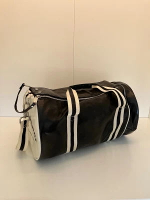 Fred Perry weekendbag - Svart weekendbag från Fred Perry med vita detaljer och breda randiga axel- och bärremmar. Väskan har dragkedja upptill, ytterficka och rund form. Perfekt för gymmet eller kortare resor. 