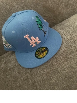 Blå LA Dodgers keps New Era 59FIFTY - Snygg blå LA Dodgers keps från New Era 59FIFTY med broderad LA-logga i ljusrosa, palmträd och patch på sidan. Flat brim och klassisk passform. Perfekt för dig som vill sticka ut med stilren streetwear.