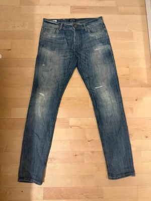 Blå slim fit jeans med slitningar - Storlek 30/34. Säljer ett par blå slim fit jeans med snygga slitningar och tvättade detaljer. Jeansen har klassisk femficksdesign, knappgylf och läderpatch bak i midjan. Perfekta för en avslappnad och trendig look.