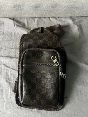 Brun axelväska med rutigt mönster - Snygg brun axelväska med klassiskt rutigt mönster och justerbar axelrem. Väskan har flera fack med dragkedja och är tillverkad i ett tåligt konstlädermaterial. Perfekt för att bära med sig det viktigaste på ett stilrent sätt.