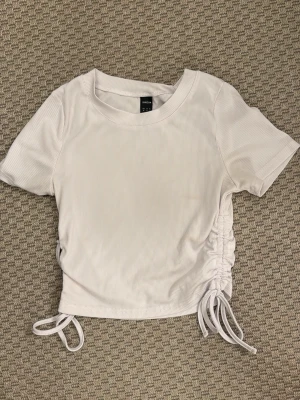 Vit ribbad topp med snörning  - Säljer en vit ribbad topp med korta ärmar och snörningsdetaljer i sidorna. Toppen är croppad och har en rund halsringning. Perfekt att styla med höga jeans eller kjol.