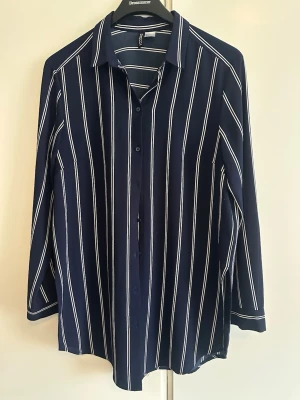 Lång Mörkblå randig blus från Divided - Längre mörkblå blus från Divided med vita vertikala ränder. Skjortan har klassisk krage, knäppning framtill och långa ärmar. Perfekt för dig som gillar en stilren och trendig look. Aldrig använd dock tvättad en gång :)