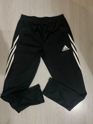 Svarta Adidas träningsbyxor - Säljer ett par svarta träningsbyxor från Adidas med de klassiska vita ränderna längs sidorna och Adidas-logga på vänster lår. Byxorna har resår i midjan och är tillverkade i ett lätt och bekvämt material, perfekta för träning eller chill.