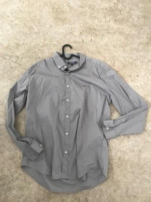 Grå skjorta från Tommy Hilfiger - Klassisk grå skjorta från Tommy Hilfiger med button-down krage och vita knappar. Skjortan har lång ärm och en liten broderad logga på bröstet. Tillverkad i bomull för en skön och stilren look.