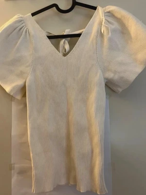 Vit ribbad topp med puffärm och knyt - Supersöt vit ribbad topp med v-ringning, puffärmar och knytband i ryggen. Toppen har en figurnära passform och är perfekt för att lyfta vilken outfit som helst. Materialet känns mjukt och stretchigt.