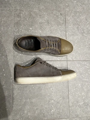 Grå sneakers från Lanvin med mocka och glansig tå - Snygga grå Lanvins med ovandel i mocka och tåparti i olivgrönt gummi. Skorna har vita sulor och matchande grå skosnören. Perfekta för dig som gillar en stilren och unik look. Lågt skaft och diskret design som passar till det mesta.