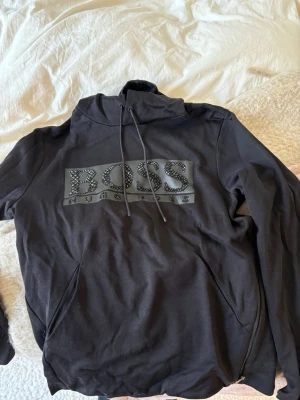 Svart hoodie från BOSS med tryck - Snygg svart hoodie från BOSS med stort logotryck framtill dekorerat med nitar. Hoodien har dragsko i huvan och praktiska fickor på sidorna. Perfekt för en chill och stilren look. Materialet känns mjukt och bekvämt mot huden.