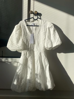 Vit kort klänning med puffärmar - Superfin vit klänning från Bubbelroom med broderade blommor,  V-ringning och stora puffärmar. Klänningen har markerad midja och knytband i ryggen. Materialet är strukturerat och ger en lyxig känsla. Perfekt för dig som gillar romantisk och trendig stil. Perfekt för student, konfirmation och skolavslutning! Ny pris 900 säljs för 800. Pris går att diskutera💕