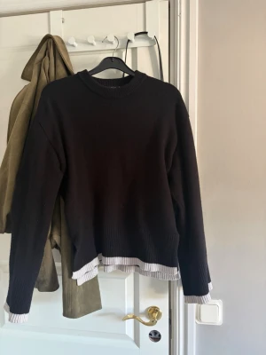Svart stickad tröja från Gina Tricot - Snygg svart stickad tröja från Gina Tricot med ribbstickade muddar och rund hals. Tröjan har en cool detalj med vit ribbad kant längst ner och vid ärmsluten. Perfekt för lager-på-lager och enkel att matcha med jeans eller kjol.