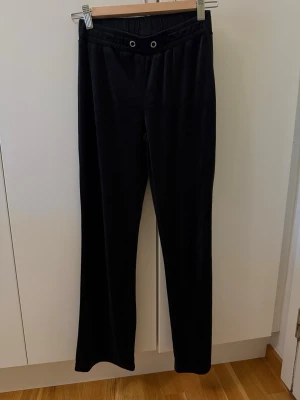 Svarta mjukisbyxor i velour - Säljer ett par svarta mjukisbyxor i mjuk velour med raka ben och resår i midjan. Byxorna har två dekorativa öljetter framtill och en enkel, clean look utan fickor. Perfekta för en chill och avslappnad stil.
