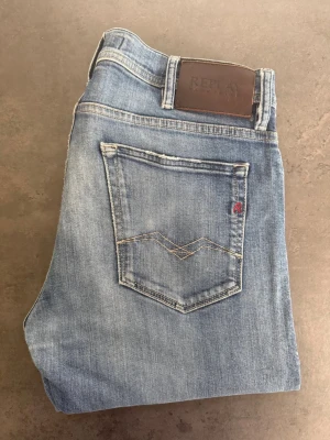 Blå slim fit jeans från Replay 33/34 - Snygga ljusblå slim fit jeans från Replay med klassisk femficksdesign och diskreta slitningar. Jeansen har en läderpatch med logga bak i midjan och dekorativa sömmar på bakfickan. Perfekta för en avslappnad och trendig look.