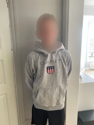 Grå hoodie från GANT med logga - Snygg grå hoodie från GANT med broderad logga och sköld på bröstet. Klassisk känguruficka framtill och huva med dragsko. Perfekt för en avslappnad och sportig stil. Mjuk bomullsblandning som är skön att ha på sig.