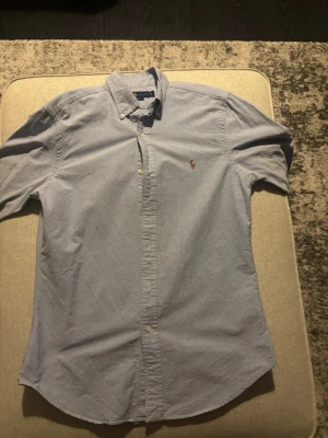 Ljusblå skjorta från Ralph Lauren - Klassisk ljusblå skjorta från Ralph Lauren med button-down krage och broderad logga på bröstet. Skjortan har långa ärmar och knäppning framtill. Perfekt för en stilren och avslappnad look.