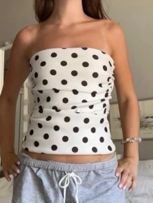 Vit prickig bandeau tubtopp - Säljer en vit bandeau tubtopp med svarta prickar. Toppen är ärmlös och har en tight passform som sitter snyggt runt kroppen. Perfekt för dig som gillar en enkel men trendig look. Materialet känns mjukt och stretchigt.
