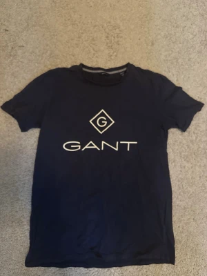 Mörkblå GANT t-shirt med logga - Snygg mörkblå t-shirt från GANT med vit logotyp och tryck på bröstet. Klassisk rund halsringning och korta ärmar. Perfekt för en stilren och avslappnad look. Storleken är 158/164. Tillverkad i mjuk bomull som känns skön mot huden.