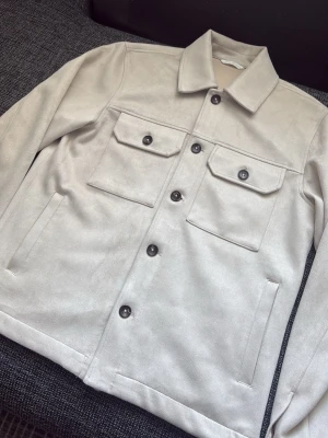Beige overshirt med fickor - Stilren beige overshirt med klassisk krage och svarta knappar. Två bröstfickor med lock och knappar samt två sidofickor. Overshirten har en rak passform och är tillverkad i ett mjukt material som påminner om mocka.