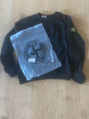 Svart sweatshirt från Stone Island - Svart sweatshirt från Stone Island med den ikoniska kompasspatchen på vänster ärm. Klassisk rund halsringning och ribbade muddar. Perfekt för en stilren och avslappnad look. Materialet känns mjukt och bekvämt.