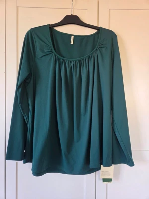 Grön långärmad topp, XL - En snygg grön långärmad topp med rundad halsringning och rynkade detaljer framtill. Modellen är lös och bekväm, perfekt för en avslappnad stil. Tillverkad i mjukt material som känns skönt mot huden.