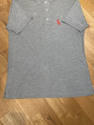 Grå pikétröja från Ralph Lauren - Klassisk grå pikétröja från Ralph Lauren med krage, knappar framtill och den ikoniska röda loggan broderad på bröstet. Tillverkad i mjuk bomull och har korta ärmar. Perfekt för en clean och stilren look.
