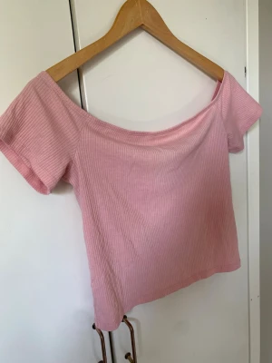 Ljusrosa ribbad offshoulder topp - Söt rosa ribbad topp med offshoulder-design och korta ärmar. Toppen är croppad och har en mjuk, stretchig känsla som gör den perfekt till jeans eller kjol. Enkel men trendig stil som passar till många olika looks.