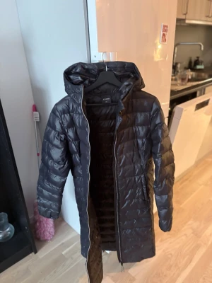 Svart lång dunjacka från Cubus - Säljer en svart lång dunjacka från Cubus med huva och dragkedja hela vägen. Jackan är quiltad och har en stilren look som håller dig varm under kalla dagar. Perfekt för vintern och enkel att matcha med olika outfits.