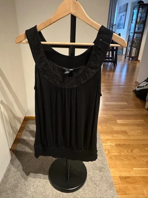 Svart ärmlös topp från H&M - Snygg svart ärmlös topp från H&M med breda axelband och rundad halsringning. Toppen har ett lätt glansigt tyg och ett löst fall, perfekt för en trendig look. Passar till många olika stilar och är enkel att matcha.