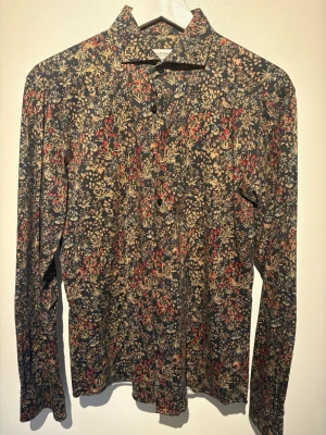 Blommig skjorta från Bruun & Stengade - Mönstrad skjorta från Bruun & Stengade i slim fit. Skjortan har ett blommigt tryck i beige, rött och blått på mörk botten, klassisk krage och knäppning framtill. Perfekt för dig som gillar unika detaljer och stil.
