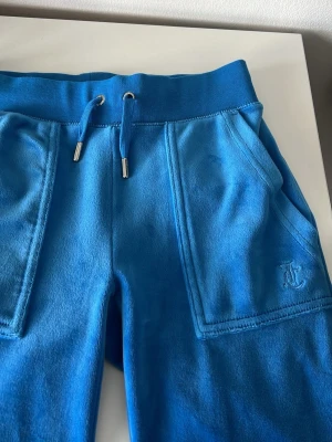 Blåa mjukisbyxor från Juicy Couture - Blåa mjukisbyxor från Juicy Couture i mjuk velour med bred resår och dragsko i midjan. Byxorna har två stora fickor fram och två bakfickor med knapp. Broderad JC-logga på ena benet. Perfekta för en chill och trendig look.