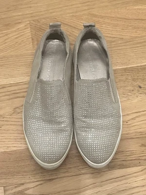 Grå slip-on sneakers med strass - Snygga grå slip-on sneakers från Tamaris med glittrande strassdetaljer över hela ovandelen. Skorna har en låg profil, vit sula och är tillverkade i mockaliknande material. Perfekta för dig som vill ha en stilren men ändå glittrig look.