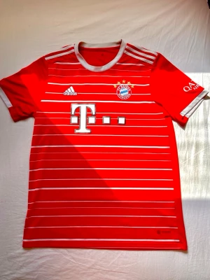 Bayern München matchtröja Coman 11 - Röd Bayern München matchtröja från Adidas med vita ränder, klubbmärke och sponsortryck fram. Nummer 11 och Coman tryckt på ryggen. Korta ärmar, rund hals och detaljer i vitt. Perfekt för fotbollsfans som vill visa sitt stöd för laget.