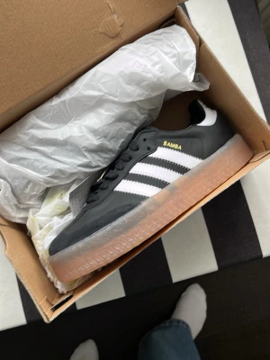 Adidas Samba OG svart/vit sneaker - Svarta Adidas sambae sneakers med mocka framtill och läder ovanpå med vita detaljer och de ikoniska tre ränderna på sidan. Gummisula i transparent brun/orange färg och guldfärgad SAMBA-text. 