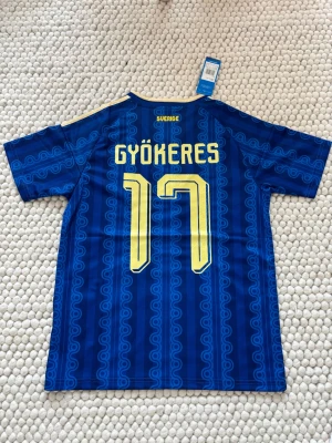 Blå Sverige fotbollströja Adidas - Säljer en blå fotbollströja från Adidas med Sveriges landslagsemblem och nummer 17 i gult. Tröjan har klassiska Adidas-ränder på axlarna, retroinspirerat mönster och v-ringad krage. Tillverkad i lätt funktionsmaterial, perfekt för träning eller match.