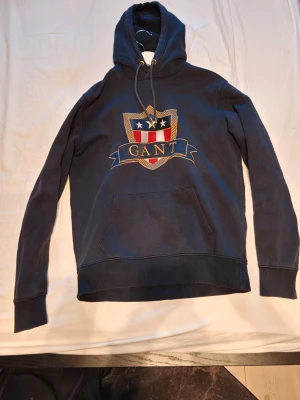 Mörkblå hoodie från GANT med emblem - Snygg mörkblå hoodie från GANT med stort broderat emblem på bröstet i rött, vitt och guld. Klassisk känguruficka framtill, ribbade muddar och justerbar huva med snören. Perfekt för en avslappnad och stilren look.