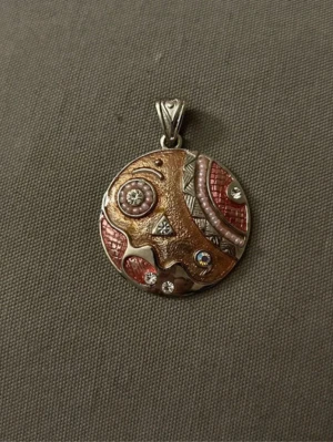 Runt hängsmycke med mönster och stenar - Unikt runt halsbandshänge i metall med abstrakt mönster i brons, rosa och silver. Dekorerat med små gnistrande stenar och pärlliknande detaljer. Perfekt för dig som gillar färgstarka accessoarer med konstnärlig touch.