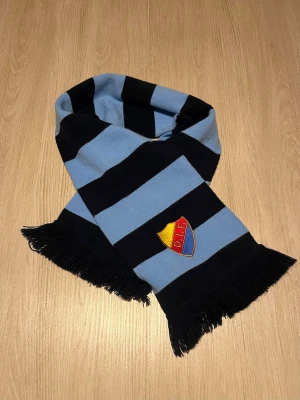 Randig Djurgården halsduk blå/svart - Snygg halsduk med breda blå och svarta ränder samt DIF:s klubbmärke broderat på ena sidan. Halsduken har fransar i ändarna och är perfekt för att visa ditt stöd på matchen. Materialet är mjukt och stickat.