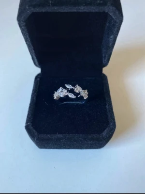 Silverring med strass stenar justerbar - Snygg silverfärgad ring med flera gnistrande, klara stenar i olika former. Ringen har en elegant och modern design där stenarna är placerade i ett mönster som påminner om löv eller blommor. Perfekt för dig som gillar stilrena accessoarer med lite extra bling.