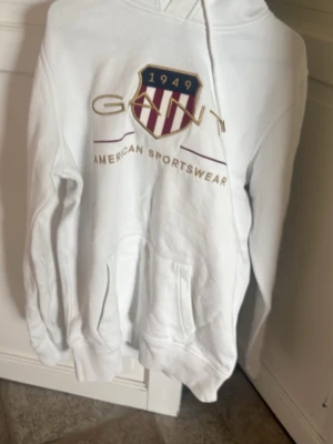 Vit hoodie från Gant med tryck - Vit hoodie från Gant med stort broderat emblem och texten 'American Sportswear' framtill. huva med dragsko. erfekt för en avslappnad look. Slutat använda det står S men passar M 