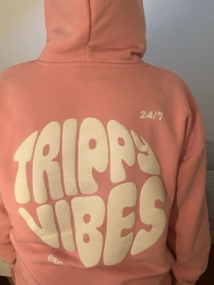 Rosa hoodie Trippy Vibes - Säljer en rosa hoodie med vit text 'Trippy Vibes' i stor retrostil på ryggen och mindre tryck framtill. Hoodien har huva med dragsko och en stor magficka. Perfekt för dig som gillar chill och snygga statement-plagg.