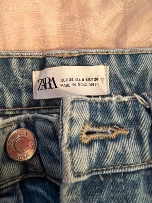 Blå raka jeans med hål från Zara - Säljer ett par ljusblå raka jeans från Zara med stora slitna hål över knäna. Jeansen har klassisk femficksdesign, knappgylf och är tillverkade i denim. Perfekta för en avslappnad och trendig look.