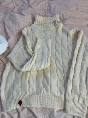 Vit kabelstickad polotröja - Kabelstickad polotröja i mjuk vit ullmix med hög krage och ribbade muddar. Snygg detalj med ett rött lönnlövsmärke nedtill. Perfekt för lager-på-lager och en clean look.